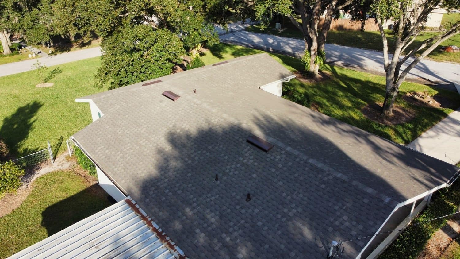 Drone view of a single-story home with a brown-gray shingle roof and box vents under large oak trees, a metal carport roof in the corner and a street behind.