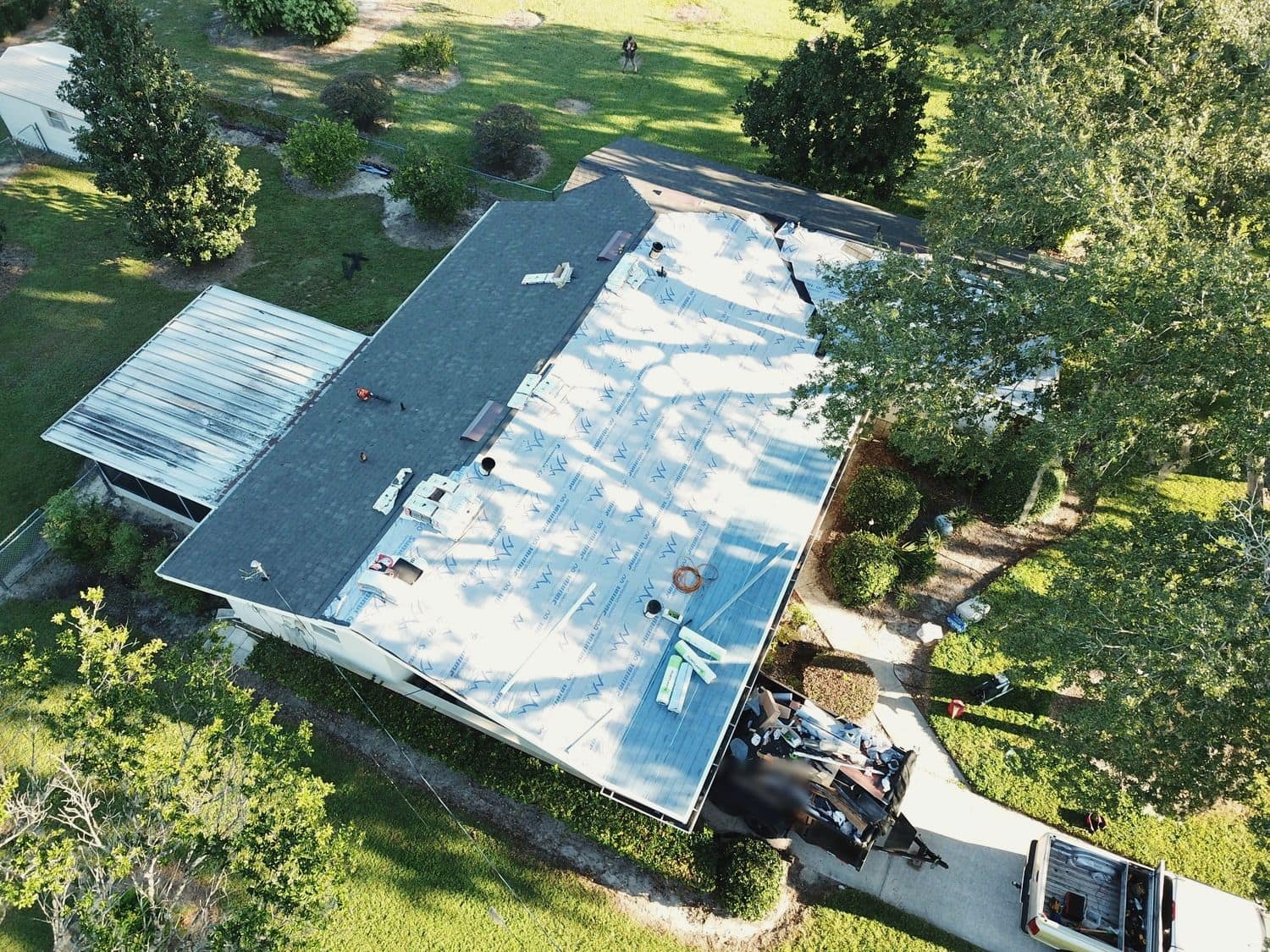 Drone view of a single-story home mid re-roof: part of the roof stripped to white synthetic underlayment, the rest still in gray shingles, a dump trailer of debris and a pickup in the driveway.
